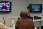 Dennis Ross, center and on left screen, a case manager for the Central Vermont Council on Aging, gives testimony to the panel. Photo by Josh Larkin.