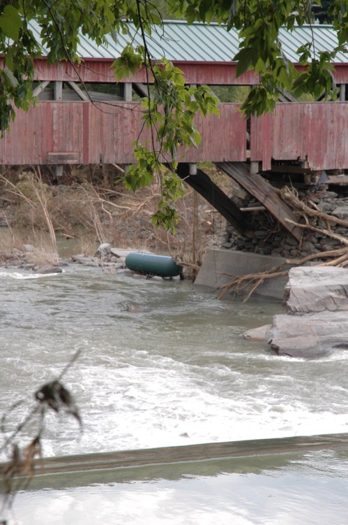 The broad span of the iconic Taftsville Covered Bridge on Rt. 4 is closed after Sunday's flood due to foundatin concerns and a large propane tanks sits in the Ottauquechee River below. VTD/Andrew Nemethy
