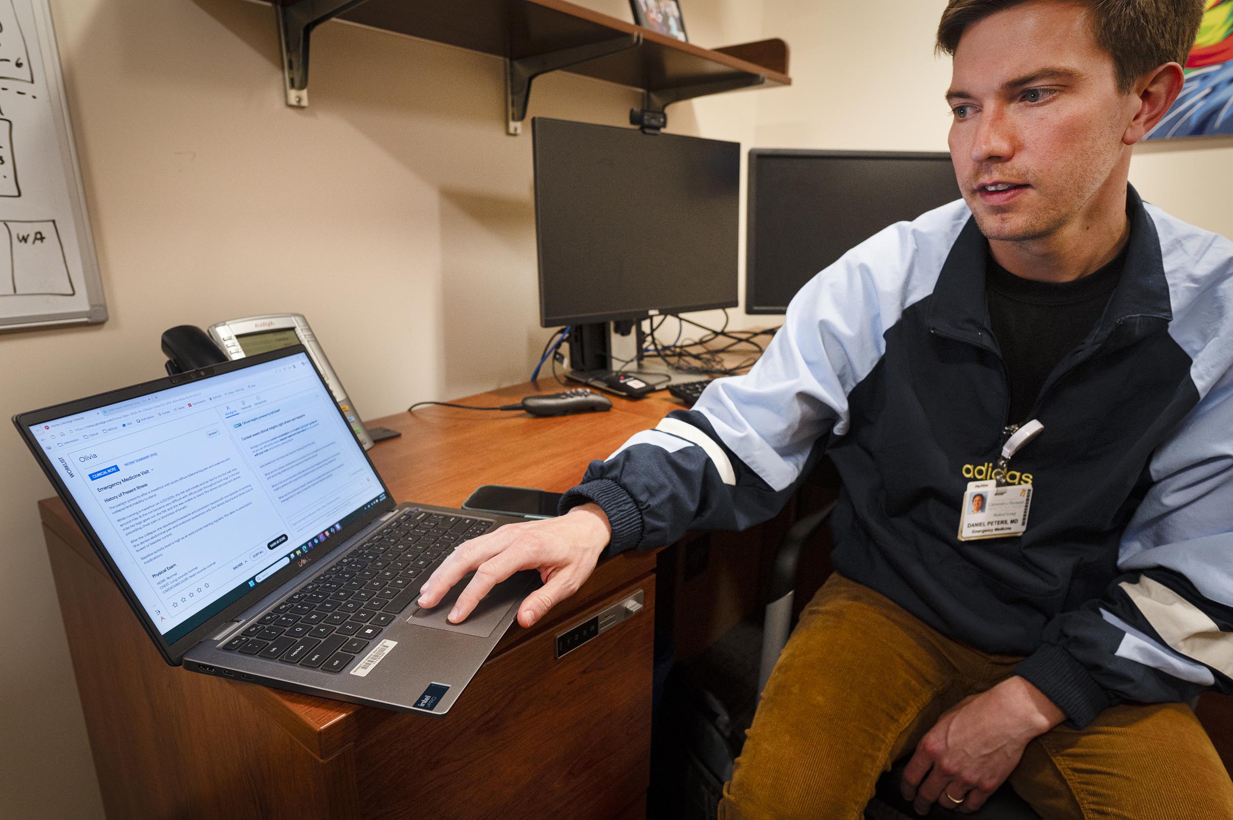A person is sitting at a desk, with two monitors and office supplies in the background, and pointing at a laptop screen with a document displayed on it.