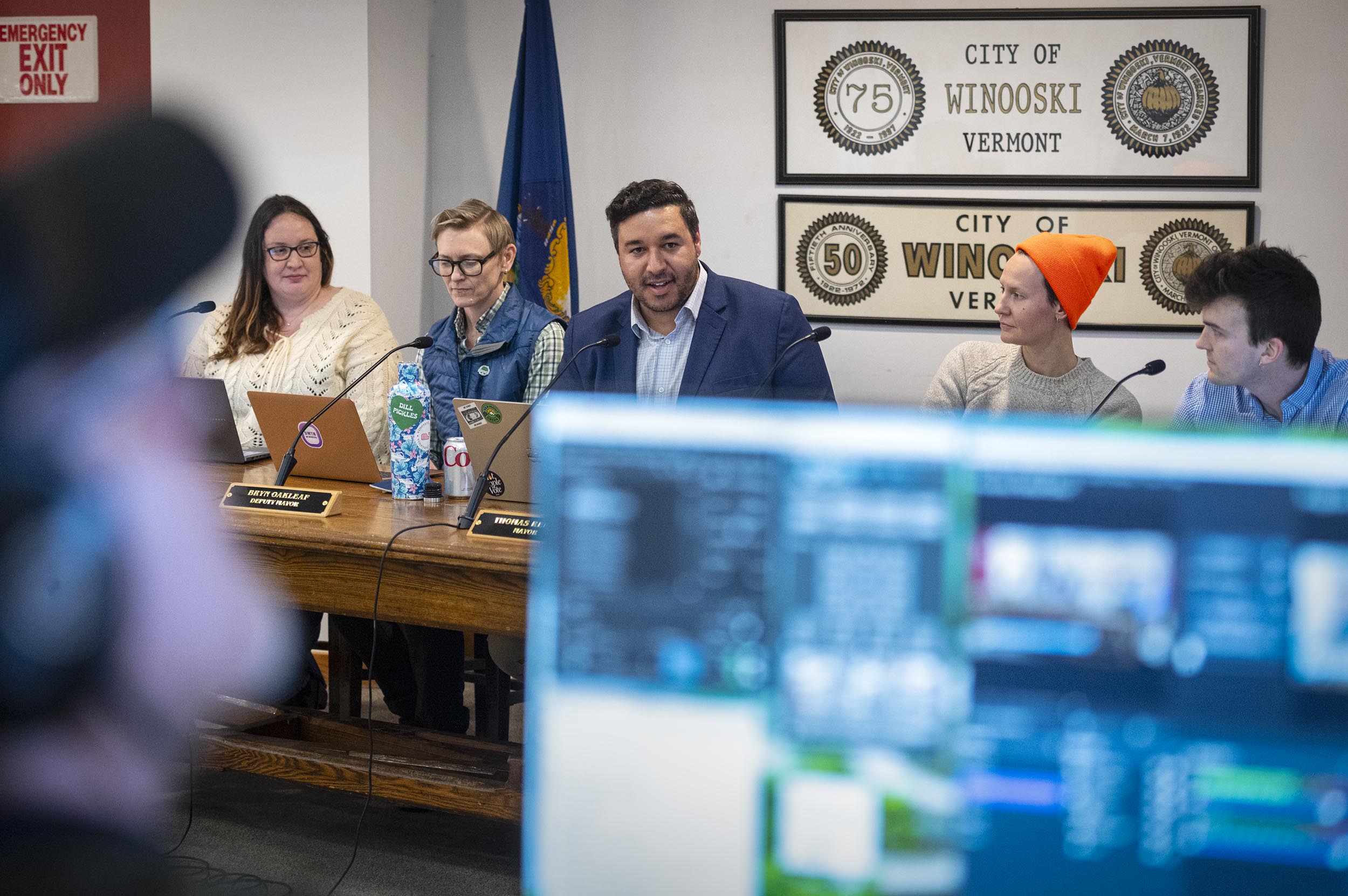 A panel of five people sits at a table during a meeting in Winooski, Vermont, with screens and audio equipment visible in the foreground.