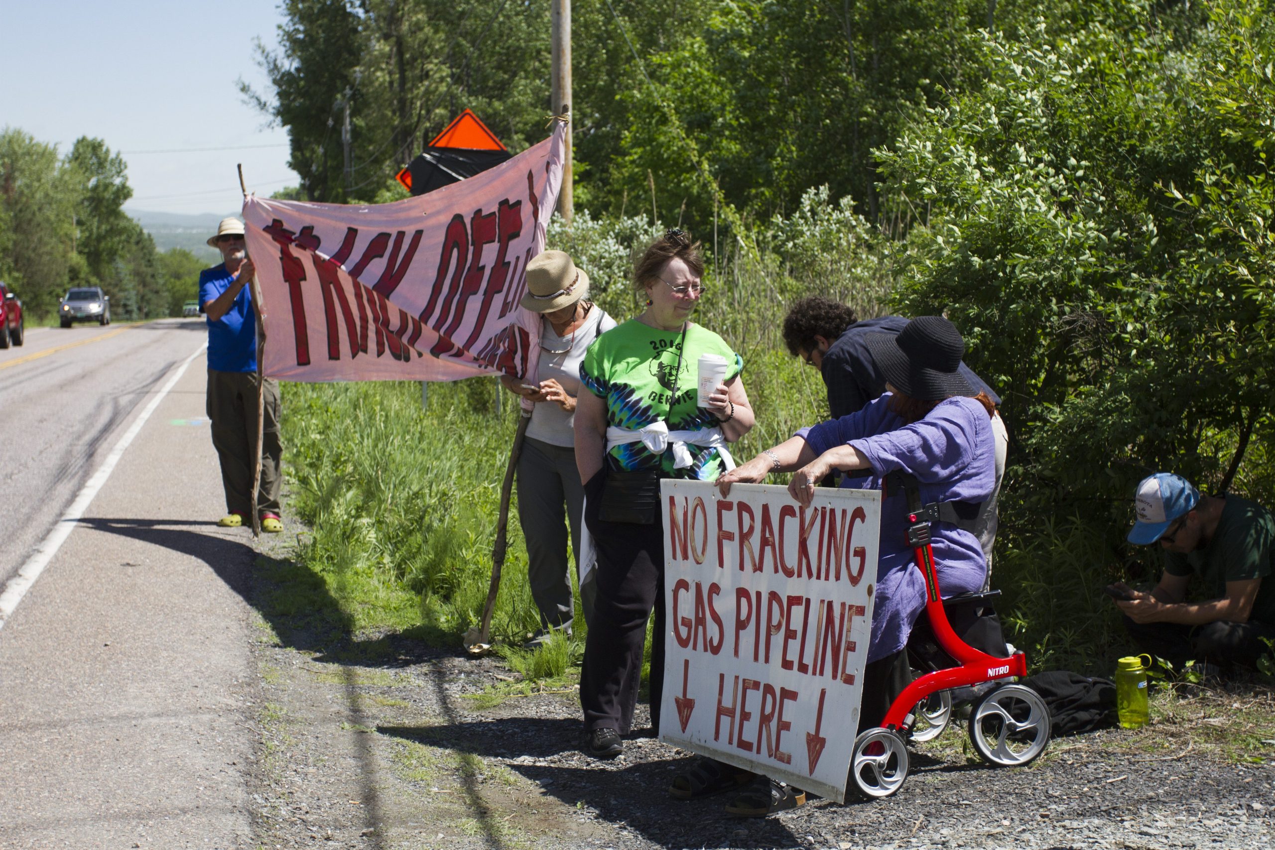 Protesters gathered near a road, holding signs against fracking and a gas pipeline, standing resolute in a grassy area. Despite the peaceful setting, some protesters were arrested as tensions heightened.