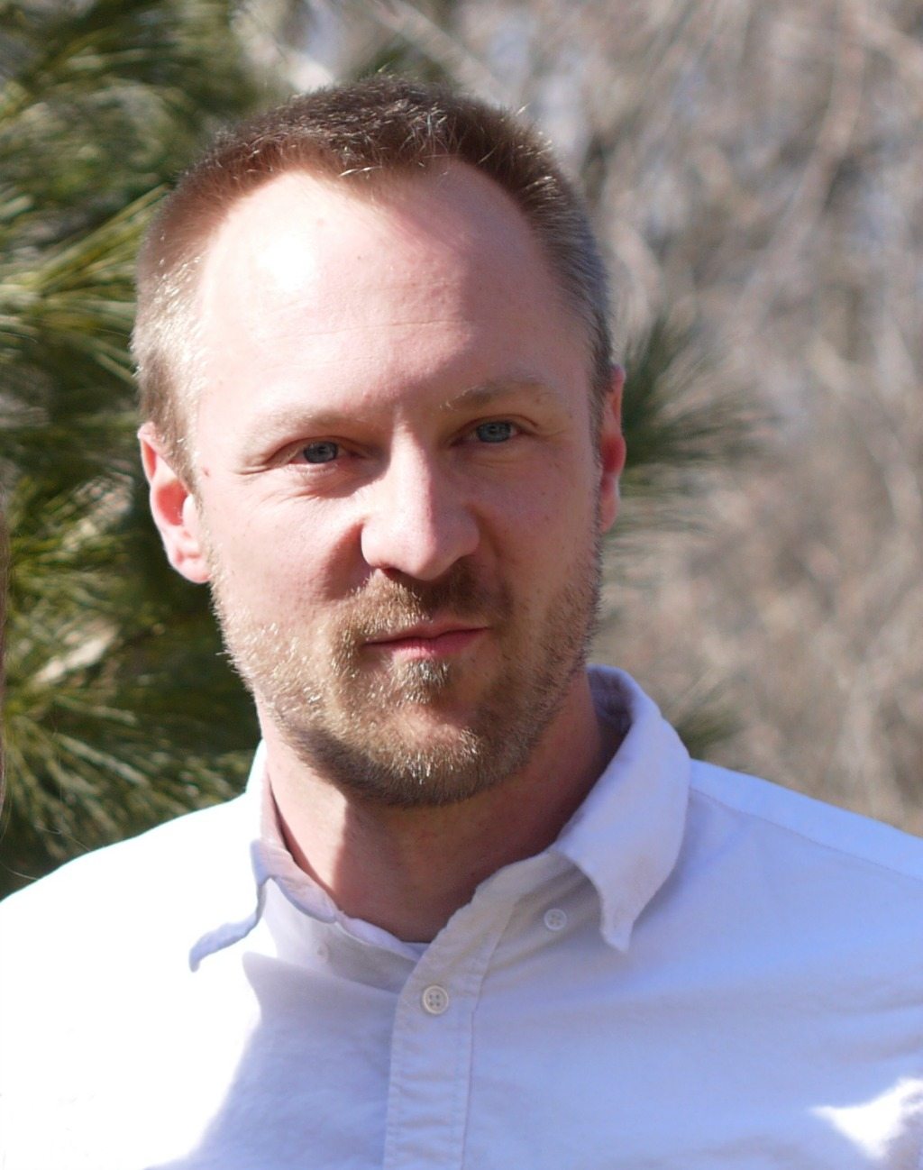 A man with short blonde hair and a beard wearing a white shirt is standing outdoors with trees in the background, perhaps discussing the upcoming primary election.