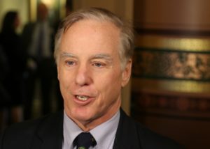 A man in a suit and tie is speaking in an indoor setting, with blurred individuals and patterned decor in the background.