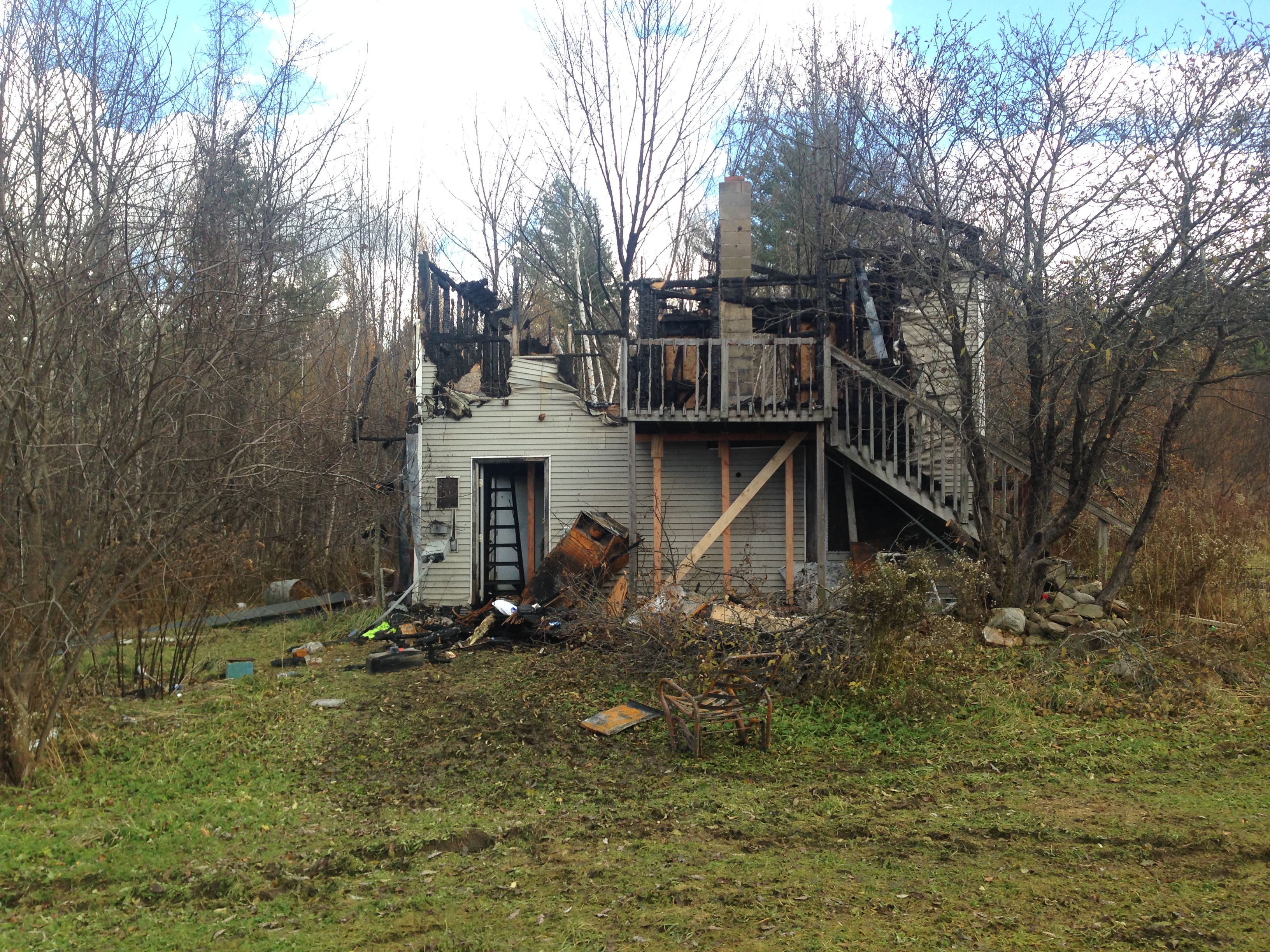 A two-story house severely damaged by fire with charred remains and debris scattered around the front yard, situated in a rural area surrounded by leafless trees.