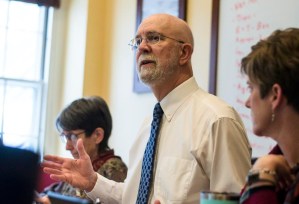A man in a white shirt and tie speaks during a meeting, with two women seated beside him; one takes notes, and a whiteboard is visible in the background.