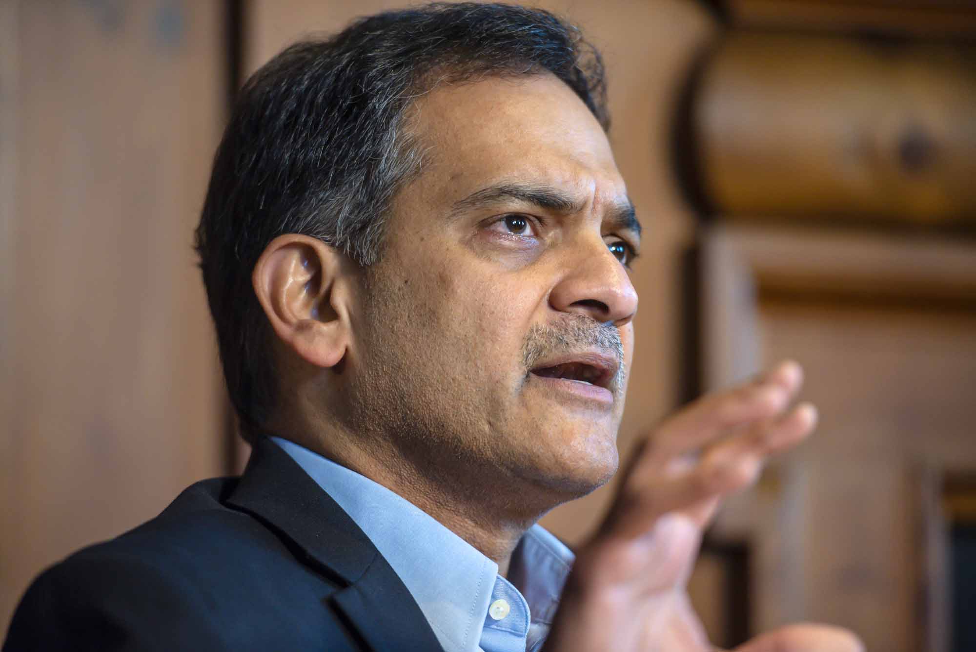 A man in a suit jacket speaks and holds his hand up.