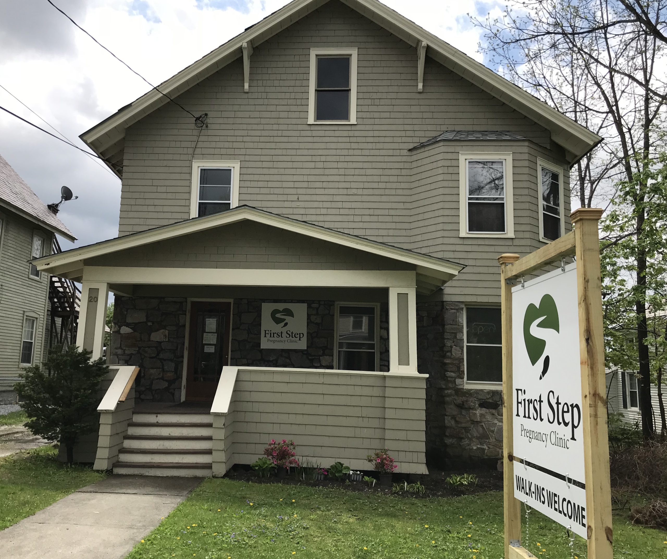 A two-story building labeled "First Step Pregnancy Clinic" with a sign by the lawn that reads "First Step Pregnancy Clinic - Walk-ins Welcome." The exterior is painted beige with stone accents.