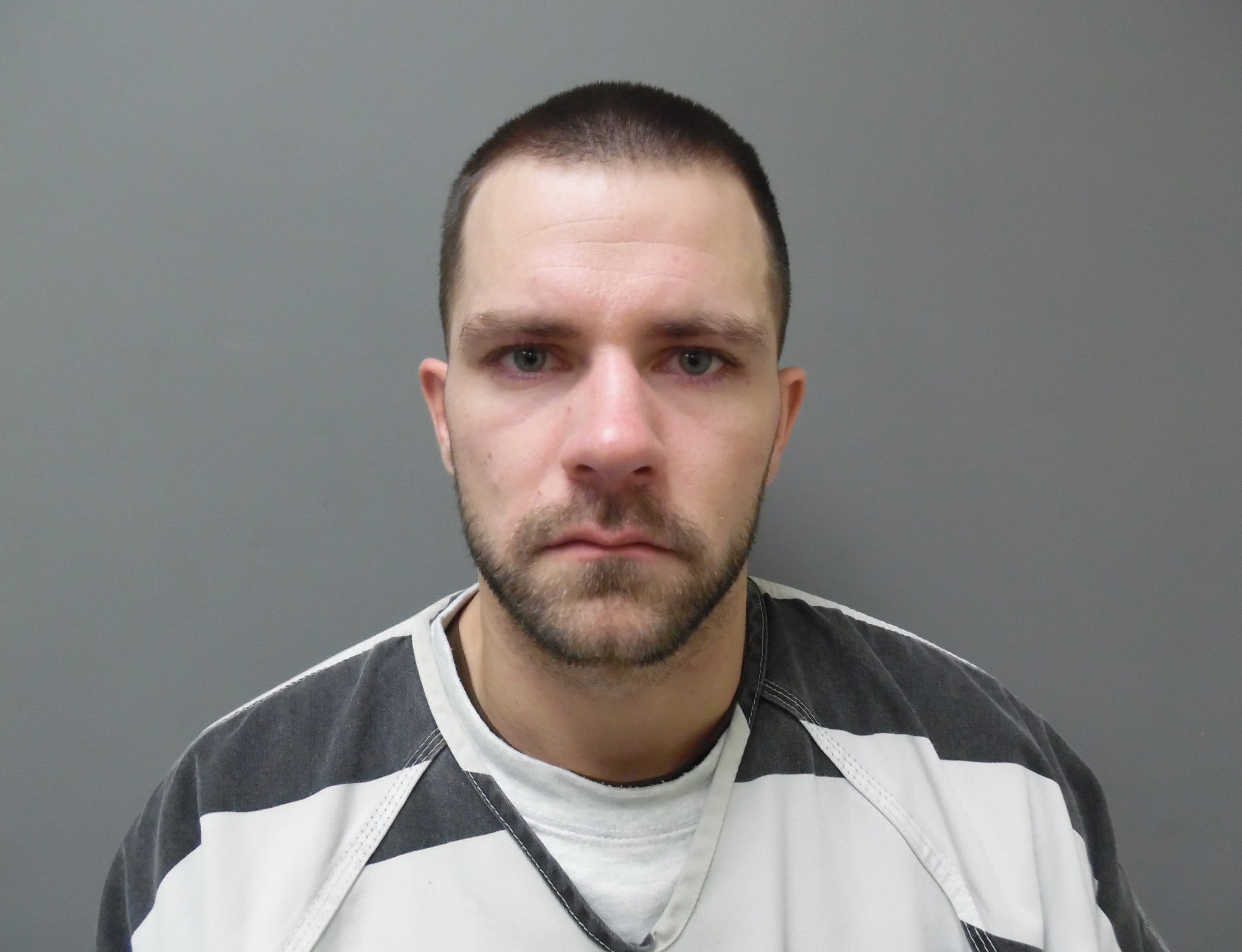 Man with short hair and light stubble, wearing a black and white striped shirt, looking directly at the camera against a grey background.