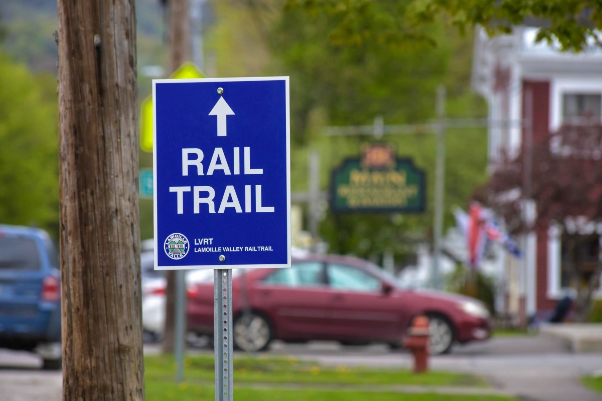 A blue sign reading "Rail Trail" with an arrow points forward; parked cars, a pole, and buildings are visible in the background.