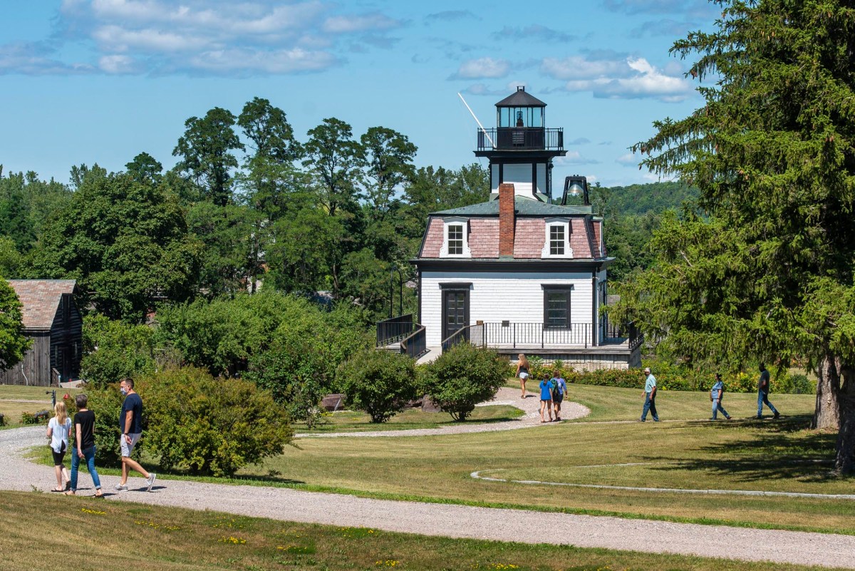 People walk along gravel paths near a white lighthouse surrounded by trees and grassy areas on a sunny day.
