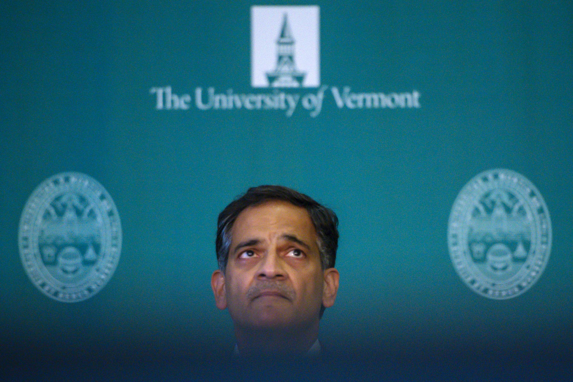 Person looking upward in front of a green backdrop that reads "The University of Vermont" with two seals on either side.