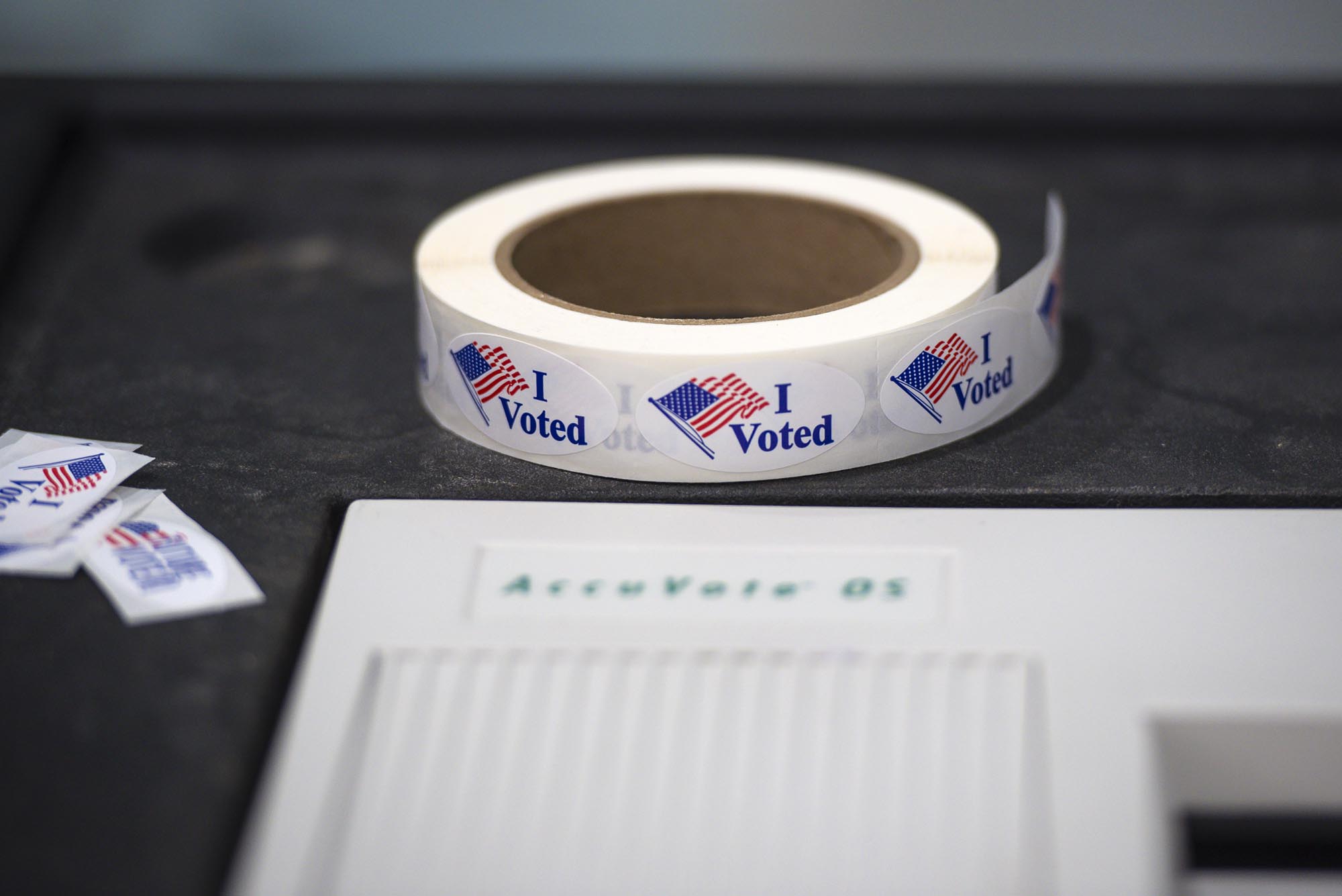 A roll of "I Voted" stickers with an American flag design is placed next to a voting machine labeled "AccuVote OS.