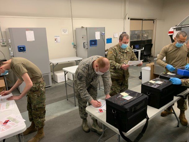 Soldiers in masks and camouflage uniforms move black containers on folding tables