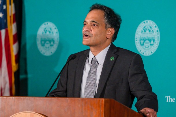 A man in a suit stands at a podium with a microphone, speaking in front of a teal backdrop featuring a circular emblem and an American flag.
