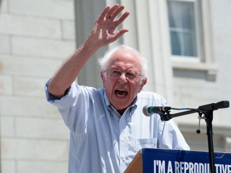 An elderly man speaks passionately at an outdoor event, raising his hand and standing behind a podium with a microphone.