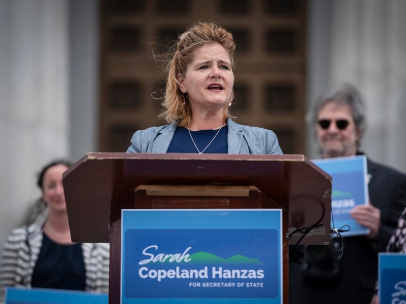 A woman speaks at a podium with a "Sarah Copeland Hanzas for Secretary of State" sign; two people holding campaign signs stand behind her.