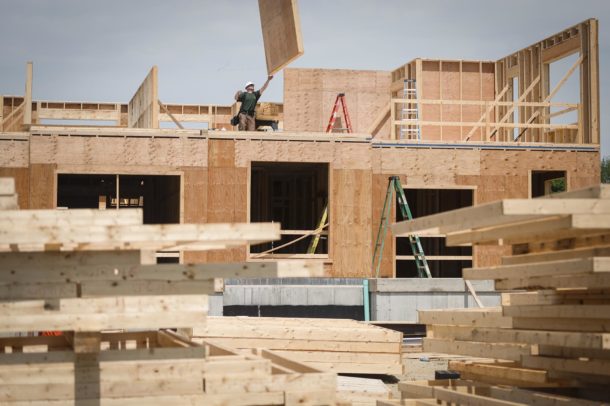 A construction worker guides a large wooden panel into place on the upper floor of a partially built wooden structure, with lumber stacks in the foreground.