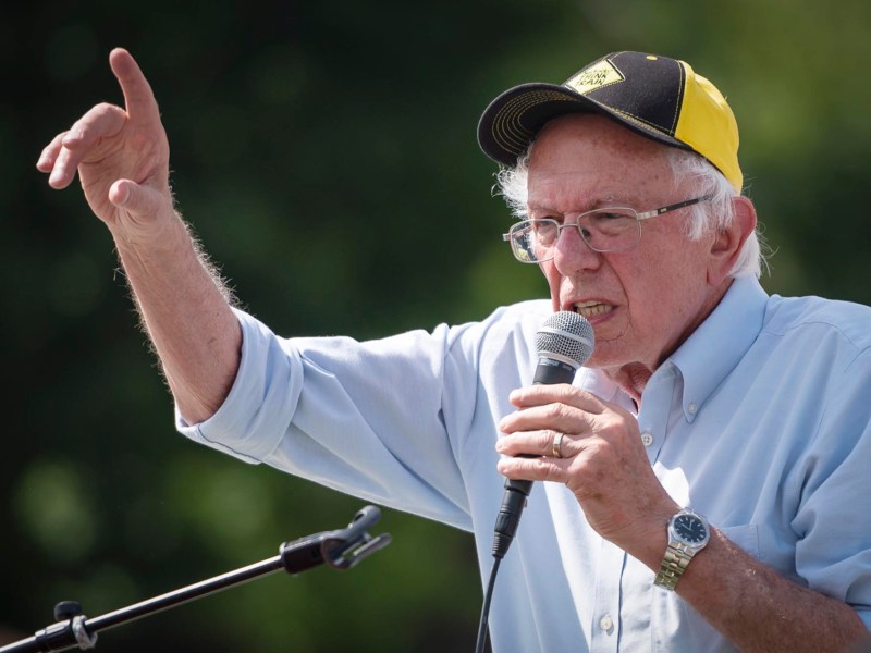 A man in a hat and blue button down shirt points his finger in the air while talking into a microphone.