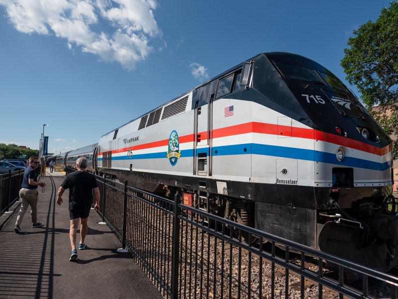 Two people walking towards a stationary train with red, white, and blue stripes on a sunny day.