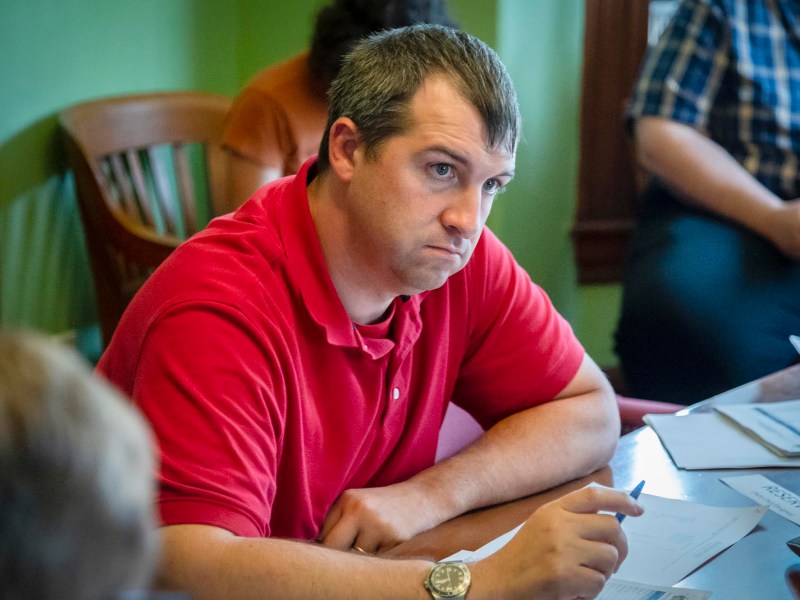 Man in a red polo shirt sitting at a table, holding a pen, looking intently forward. Papers and other individuals can be seen around him.