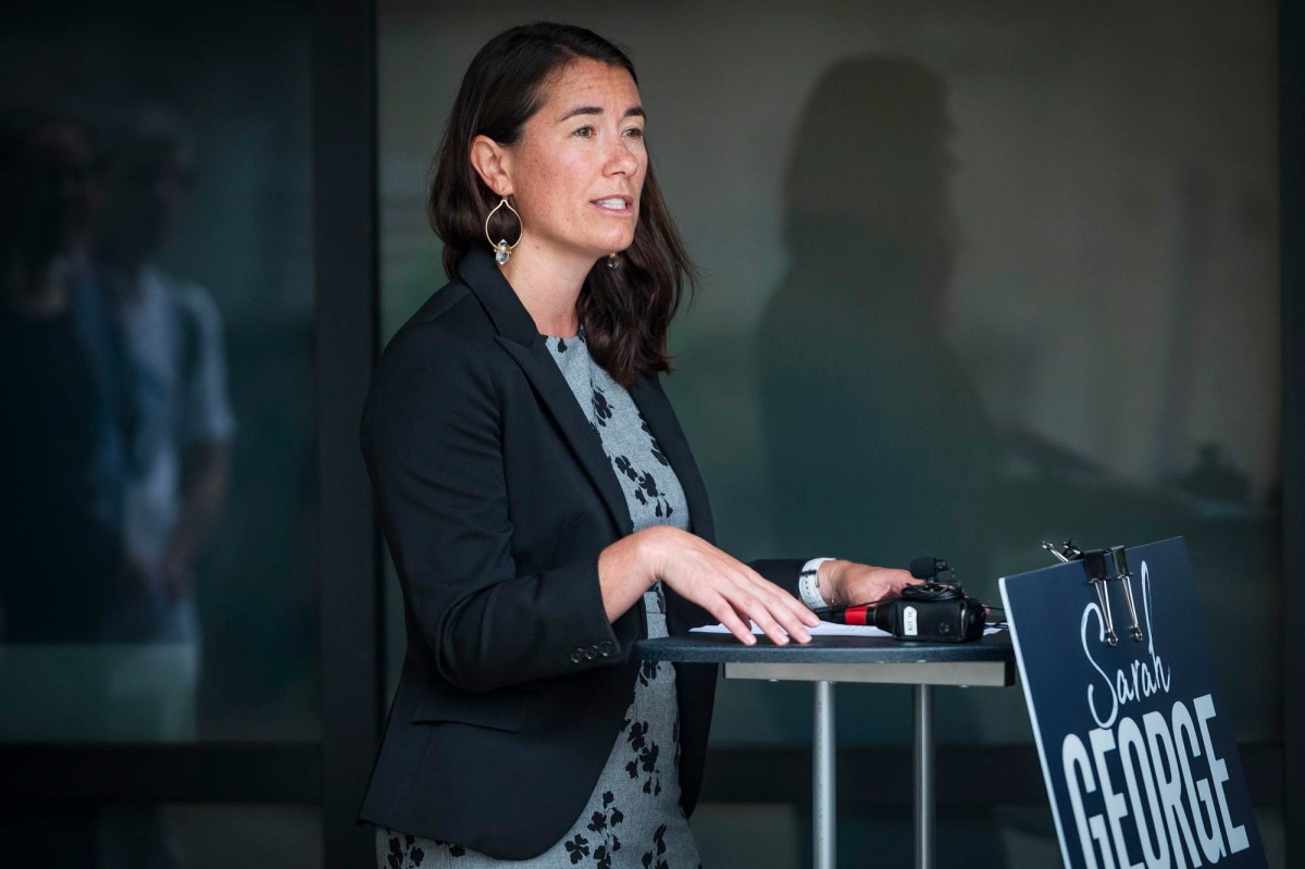 A woman in a blazer speaks at a podium with a sign reading "Sarah George" attached to it.