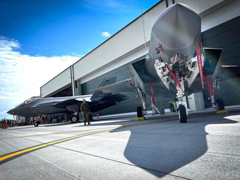 Two fighter jets are parked outside a hangar on a sunny day, with a few people and a soldier standing nearby.