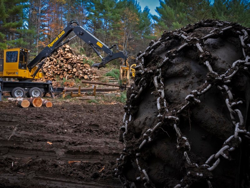 A heavy-duty logging vehicle loads logs in a forested area, with a close-up of a tractor tire covered in mud and chains in the foreground.