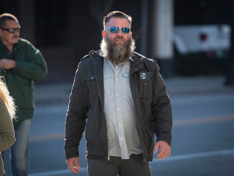 A bearded man wearing sunglasses and a dark jacket walks on a street with two other people in the background.