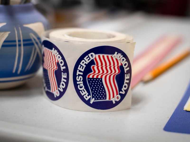 A roll of "Registered to Vote" stickers placed on a table next to a pencil, a vase, and some papers.