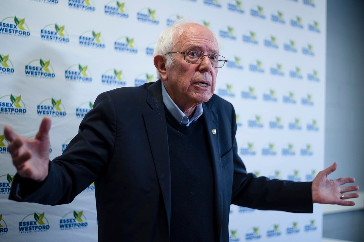 An older man with white hair and glasses, wearing a dark blazer and sweater, gestures with open arms in front of an "Essex Westford" backdrop.