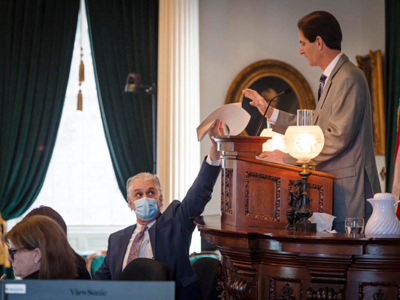 A seated man in a mask reaches toward a standing man holding papers in a room with green curtains and a U.S. flag.