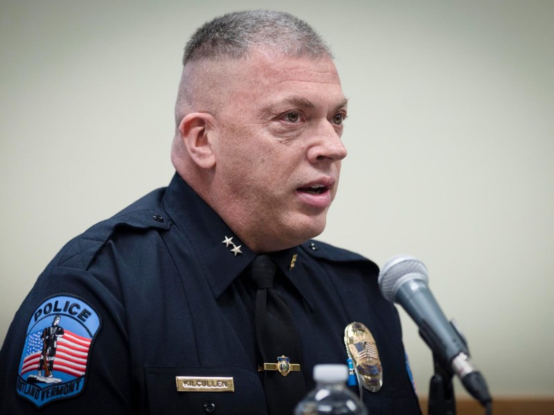 A police officer in uniform speaks into a microphone at a desk, with a badge and American flag patch visible on his shirt.