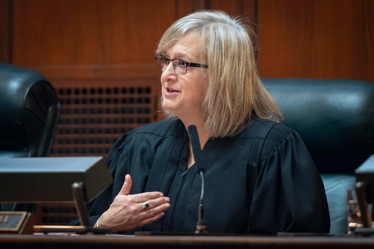 A judge in a black robe speaks, gesturing with her left hand, seated in a courtroom setting with wood paneling and a microphone.