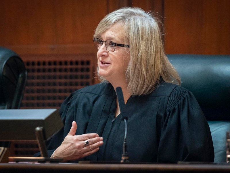 A judge in a black robe speaks, gesturing with her left hand, seated in a courtroom setting with wood paneling and a microphone.