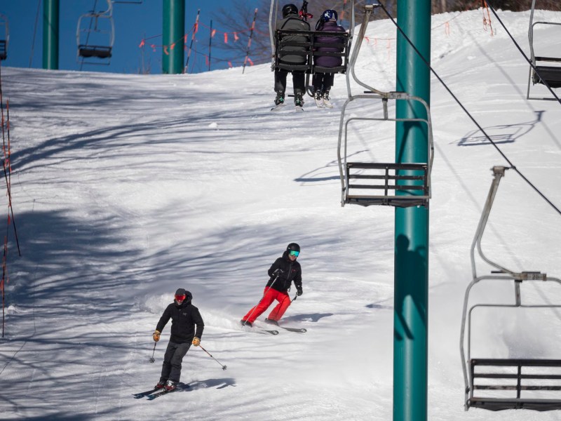 Two skiers in motion on a snowy slope under a blue ski lift carrying three people.