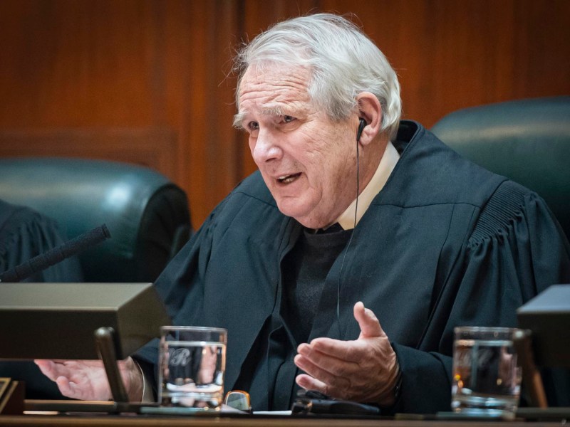 An elderly man in a judicial robe speaks while seated at a bench, with microphones and glasses of water in front of him.
