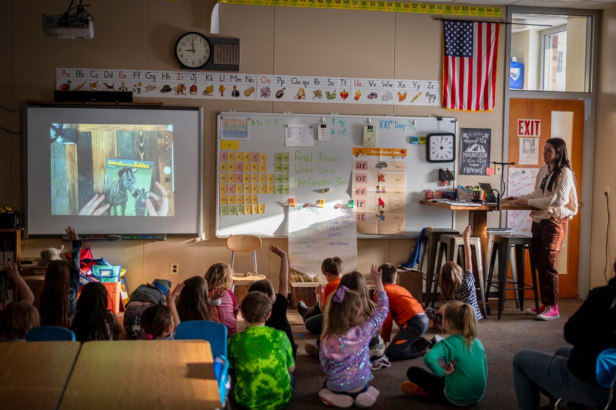 A teacher stands at the front of a classroom pointing at a projection screen, while children sit on the floor and at desks, some raising their hands. The American flag and educational materials are visible.