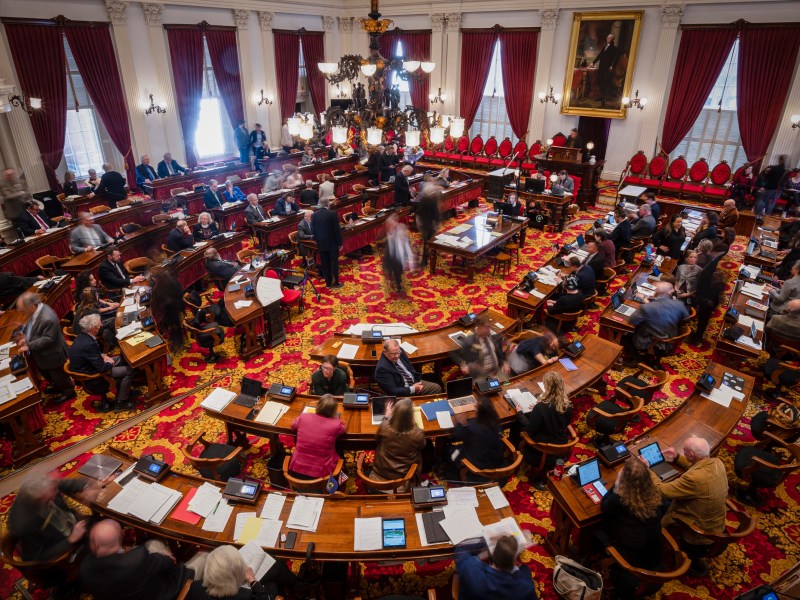 A legislative assembly in session, with members seated at desks arranged in a semi-circle inside a large, ornate chamber with red curtains, wooden furniture, and a chandelier hanging from the ceiling.