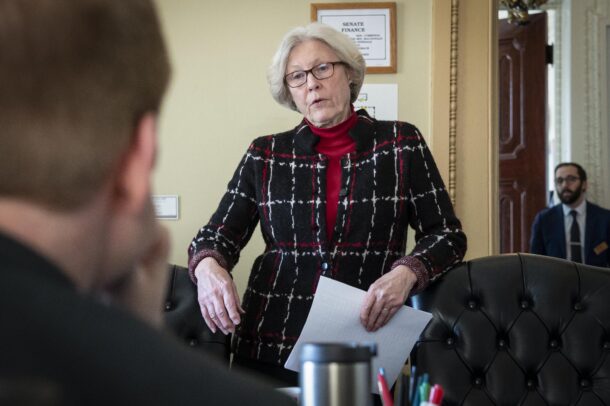A woman in a black and red checkered jacket and red scarf stands talking in an office setting, with listeners partially visible.