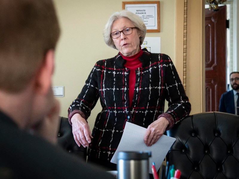 A woman in a black and red checkered jacket and red scarf stands talking in an office setting, with listeners partially visible.