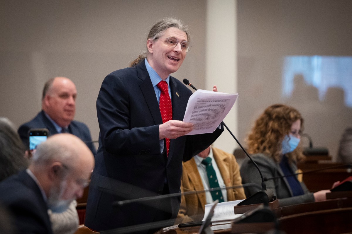 A man in a suit and red tie stands speaking and holding papers at a legislative session, with other attendees seated around him.