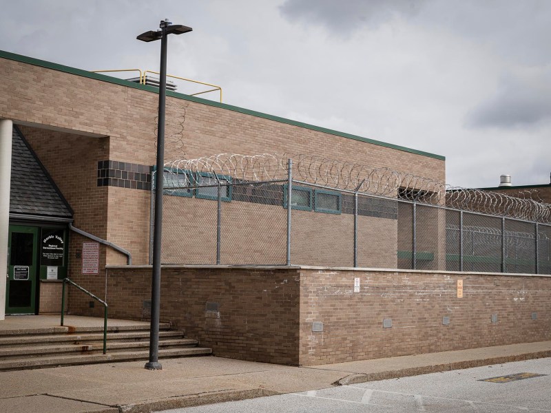 Exterior view of a brown brick building with a barbed wire fence, under an overcast sky.