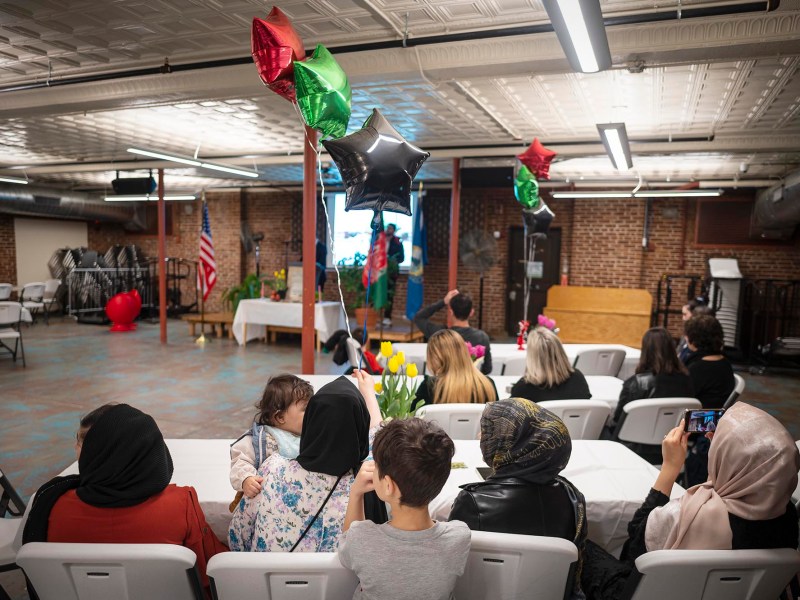 People sit at tables decorated with balloons and flowers, facing a screen in a room with brick walls and an American flag in the background.
