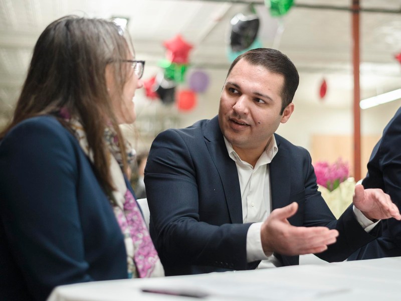A man in a suit is talking to a woman in a suit who is wearing glasses. They are seated at a table with a white tablecloth. The background is decorated with colorful hanging ornaments.
