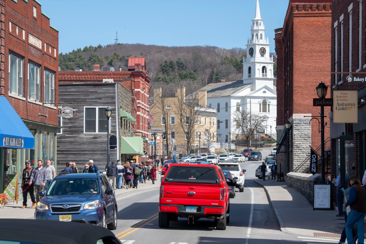 A busy street scene in a small town with pedestrians and vehicles, featuring a prominent church steeple in the background.