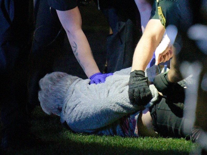 Police officers detain a person on the ground at night, with a bystander looking on in the background.