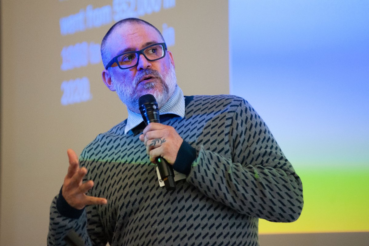 A man in a gray sweater speaks into a microphone in front of a project slideshow.