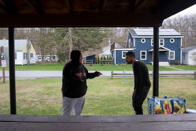 Two people have a conversation outside on a grassy area with houses in the background on an overcast day. One person wears a hoodie and the other a zipper jacket. A colorful shopping bag is on the ground.