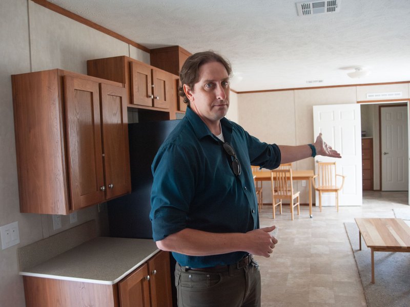 A man in a blue button-down gestures toward a living space in a manufactured home.