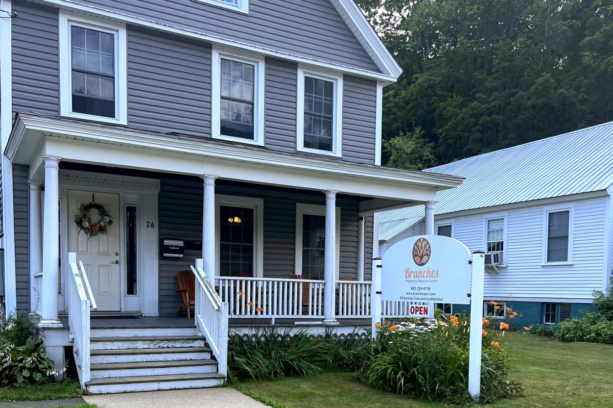 A gray and white house with a porch, a sign reading "Branches," and a small "OPEN" sign. The house is surrounded by greenery and a white neighboring house is visible in the background.